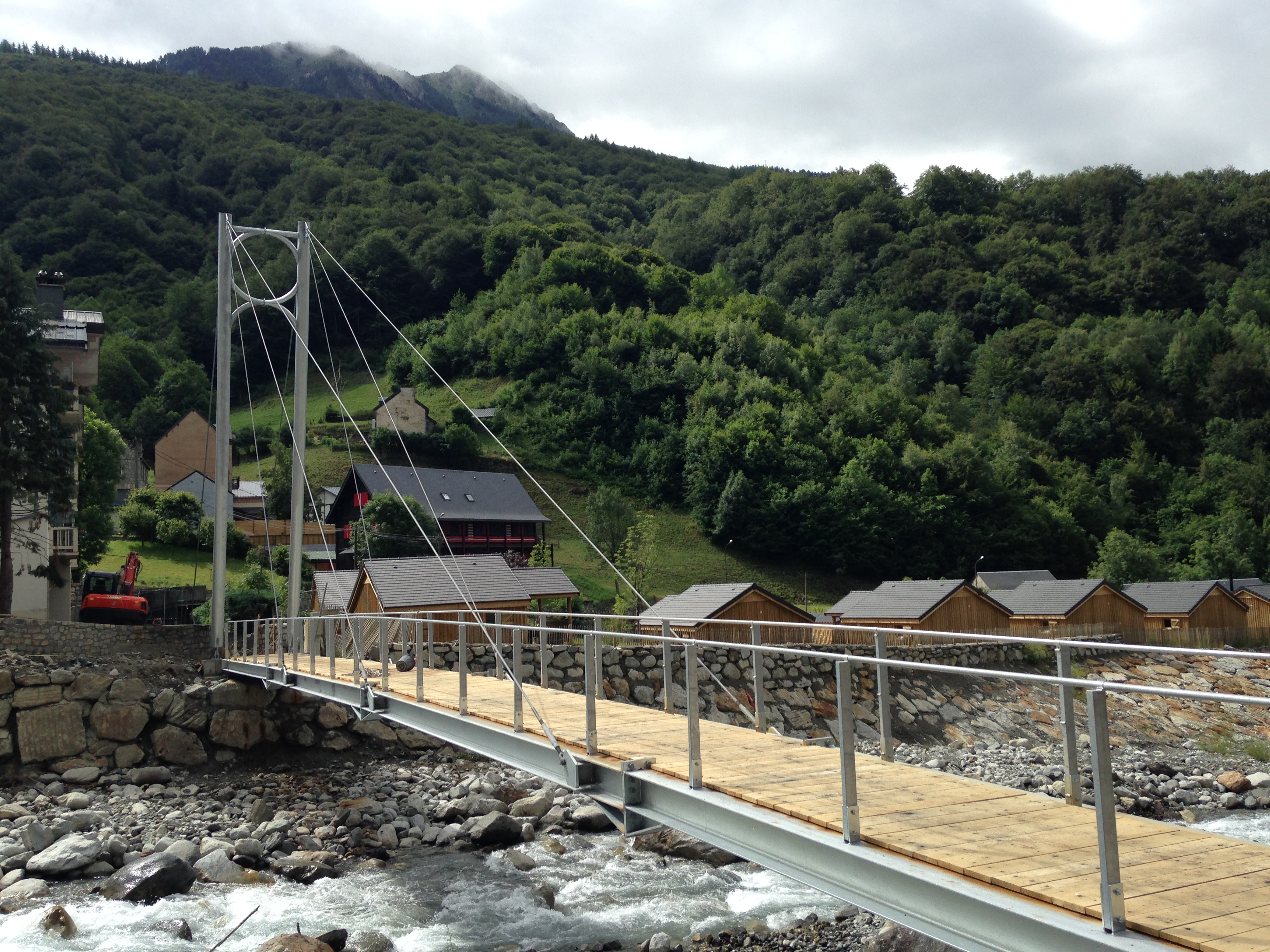 Vue d'une passerelle suspendue traversant une rivière en zone montagneuse, intégrée dans un village rural